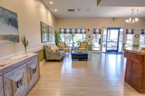 Bright senior living lounge with seating, plants, artwork, and a reception desk.