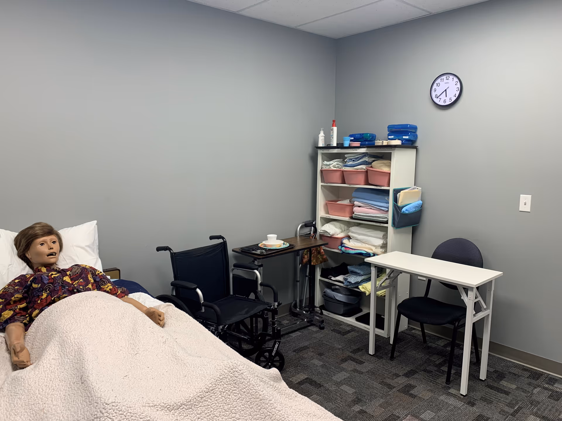 Clinical room with a bed, mannequin under a blanket, wheelchair, and towel shelf.