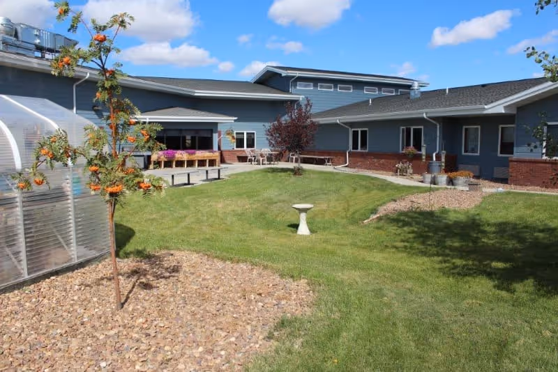 Outdoor view of Prairie Ridge Village LLP showing a green lawn with a small tree and a birdbath in the center. The building has blue siding with brick accents and multiple windows. There is a clear greenhouse structure on the left side and a patio area with chairs and flower boxes near the building. The sky is blue with some clouds.