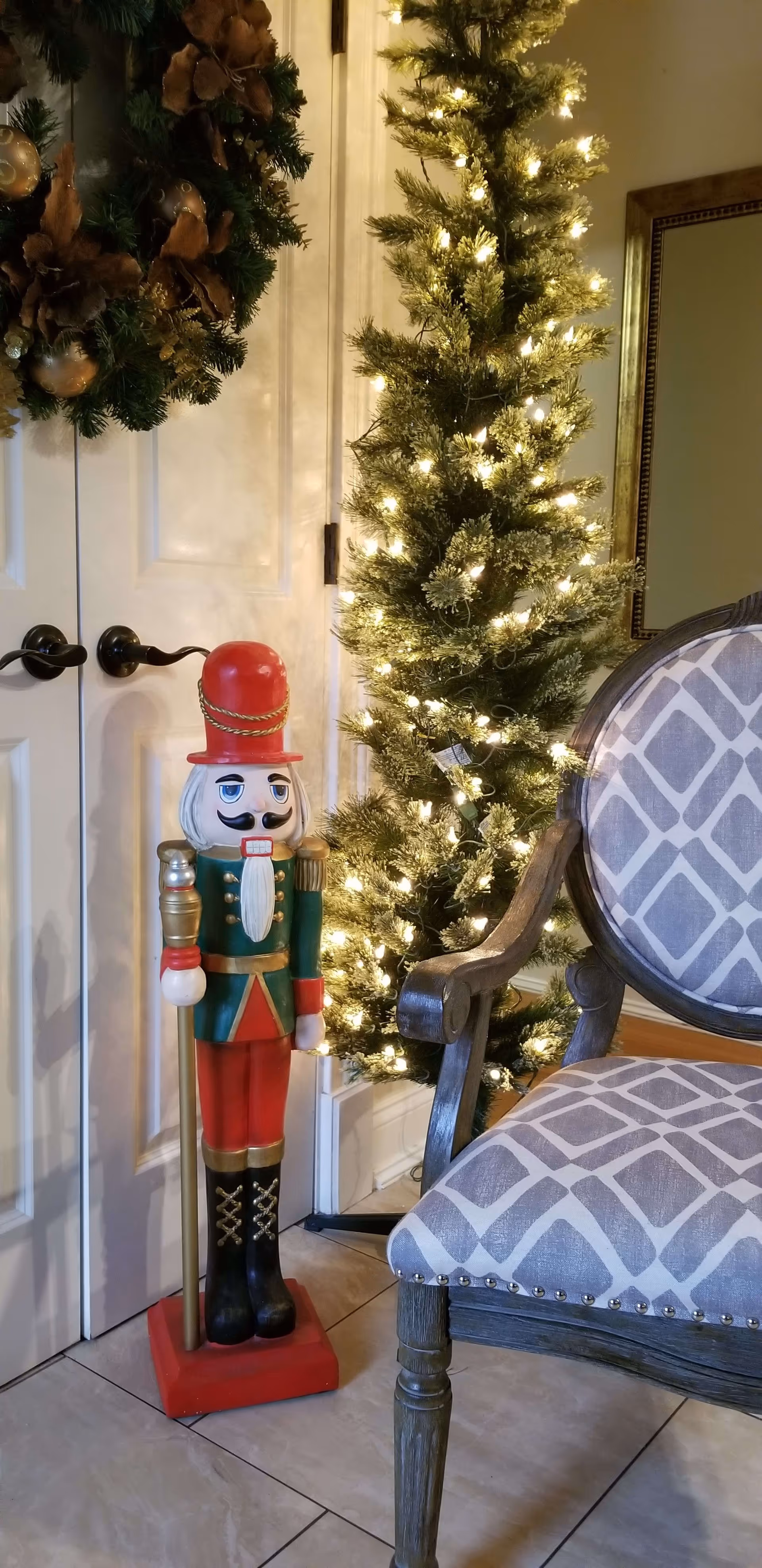 A festive indoor scene featuring a tall, lit Christmas tree next to a white double door adorned with a green wreath. In front of the door stands a decorative nutcracker figure dressed in a red hat and uniform. To the right, there is a wooden chair with a patterned gray and white cushion.