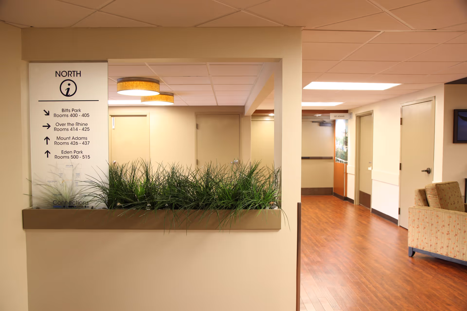 Nursing home interior hallway with planter, directional sign, doors, and seating.