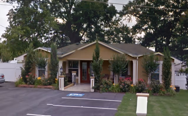Single-story beige senior care home front with red door, landscaping, and handicapped parking.