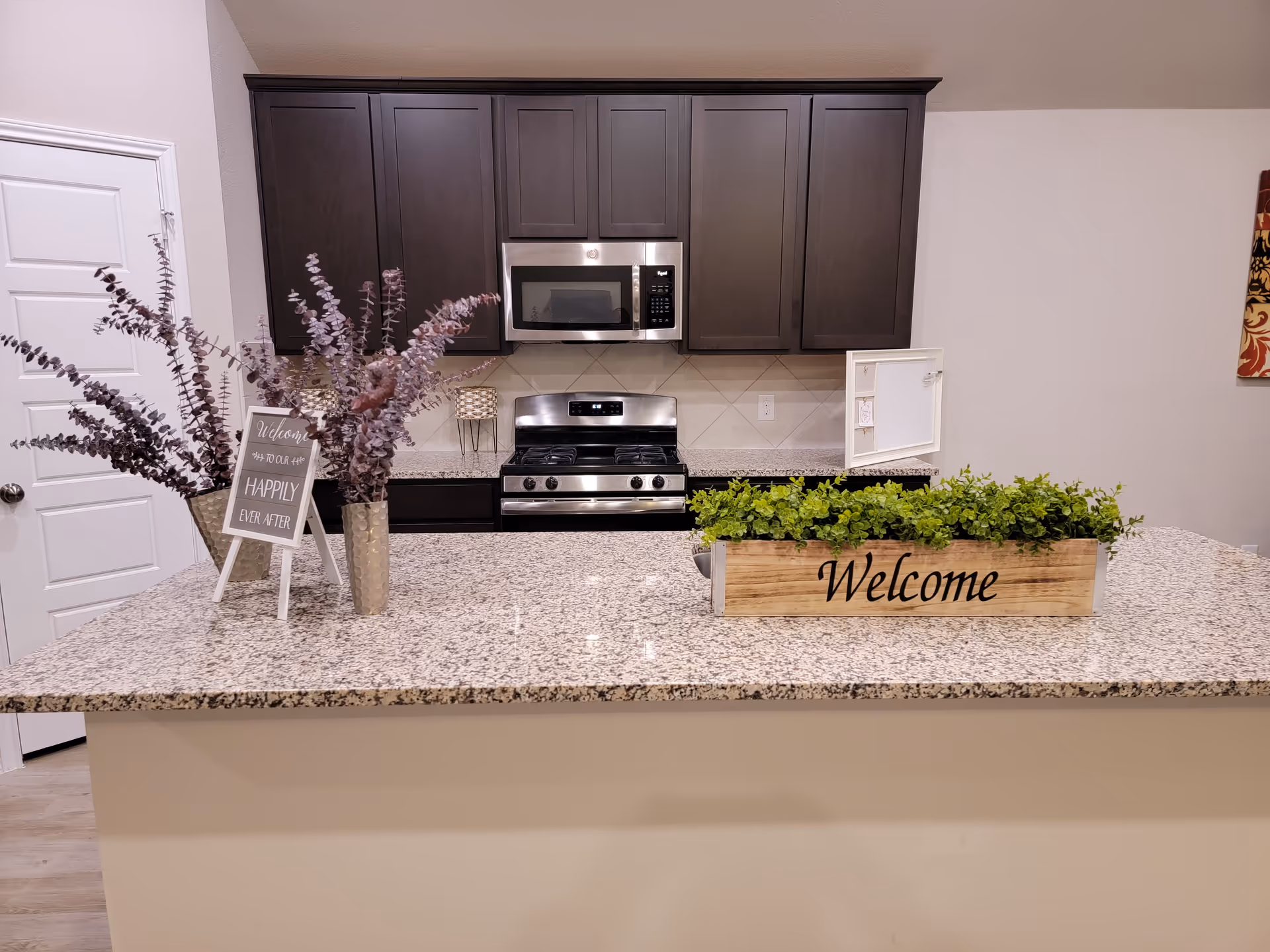 Modern kitchen with granite island, dark cabinets, stainless appliances, and a welcome planter.