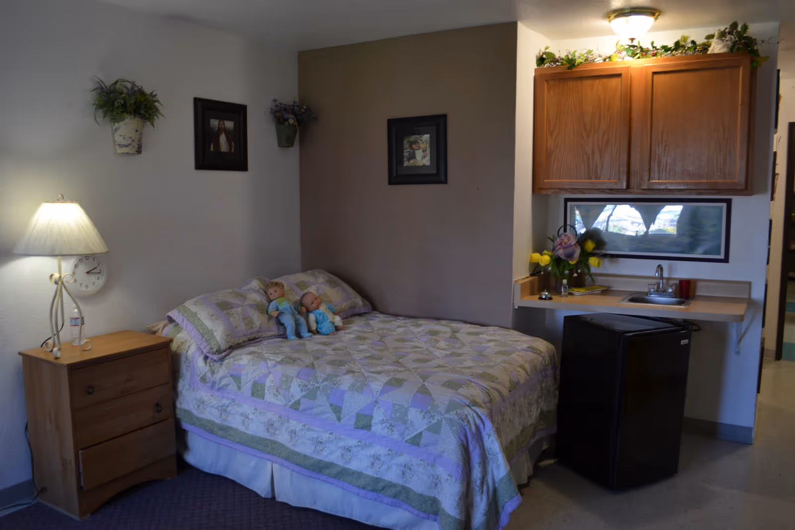 A cozy bedroom with a bed covered in a pastel quilt and two dolls sitting on it. To the left of the bed is a wooden nightstand with a lamp and a clock on the wall above it. On the right side, there is a small kitchenette area with a sink, wooden cabinets, a mini fridge, and a vase with flowers. The walls have framed pictures and decorative plants.