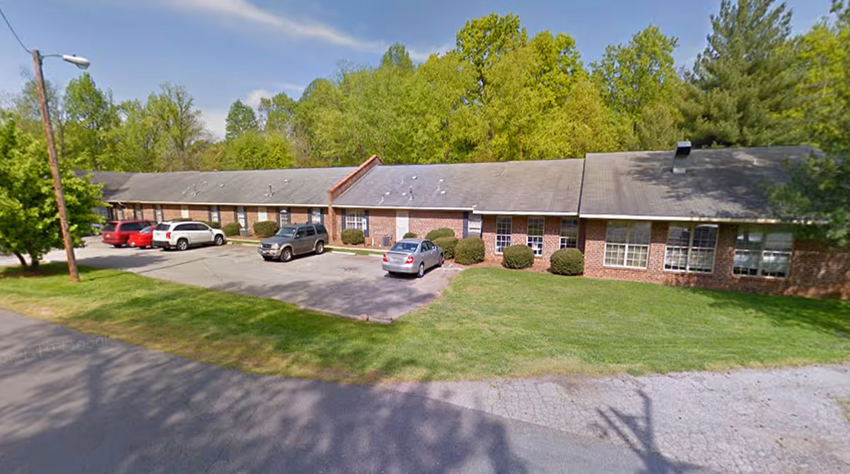 Front view of a single-story brick long-term care building with a small parking lot, several parked cars, and trees behind it.