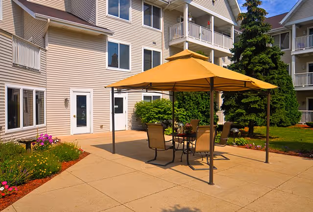 Outdoor patio area at ARIA at Mitchell Manor with a yellow canopy covering a table and four chairs, surrounded by greenery and flowers, adjacent to a multi-story residential building with balconies and windows.