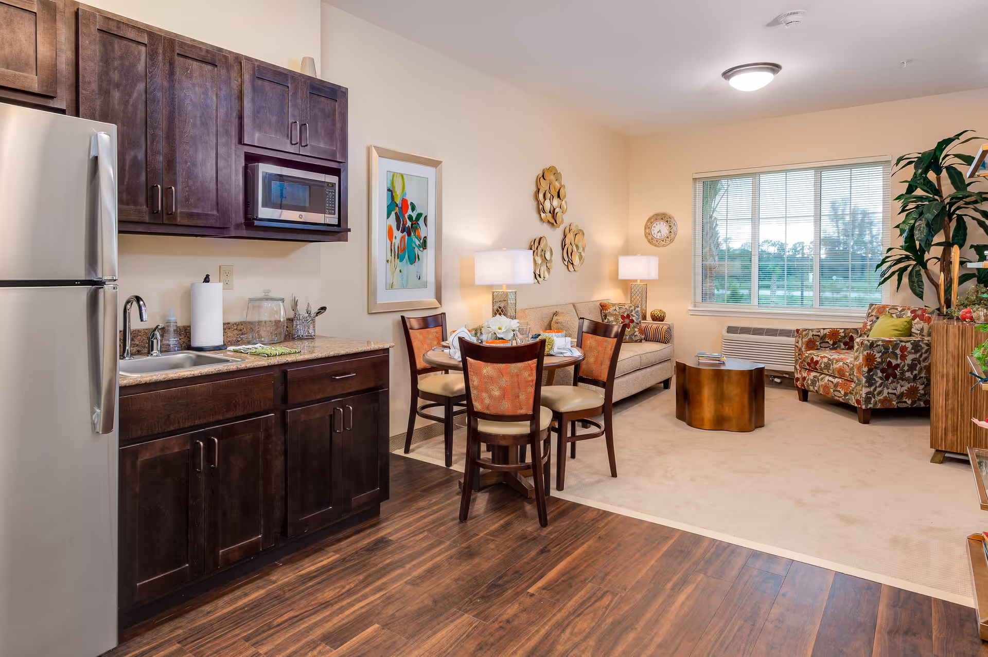 A cozy living space featuring a small kitchen area with dark wooden cabinets, a stainless steel refrigerator, microwave, and sink. Adjacent to the kitchen is a round dining table with four chairs. The living area includes a beige sofa, a floral armchair, a round wooden coffee table, two table lamps, wall art, a clock, and a large window with blinds overlooking greenery outside.