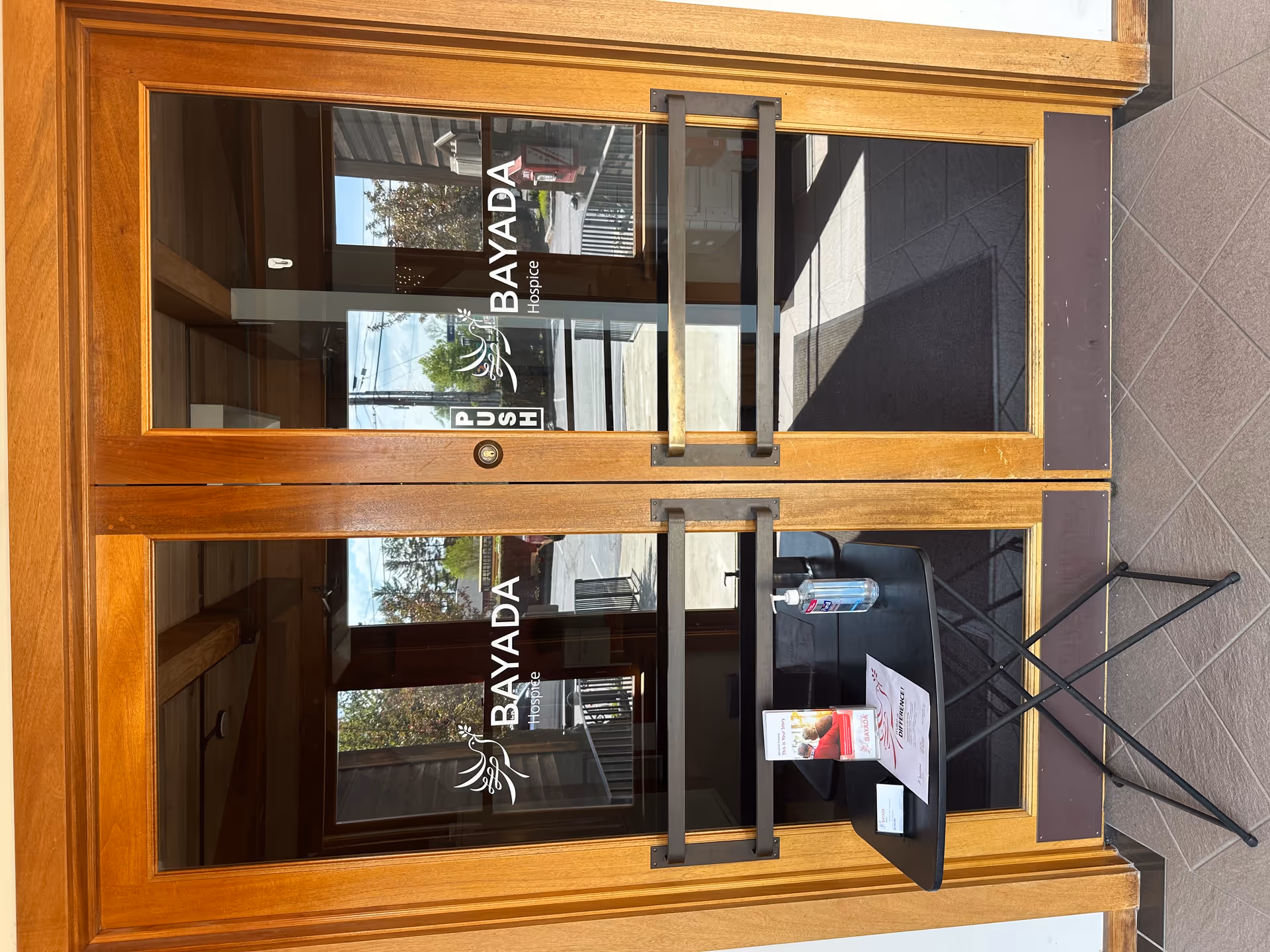 Wood-framed glass doors with BAYADA Hospice logo; outdoor tiled entry, small black table with sanitizer.