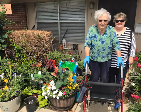 An elderly woman using a walker and wearing blue gloves stands outside a residential unit with a younger woman behind her. They are surrounded by various potted plants and flowers in front of a window and door of the building.