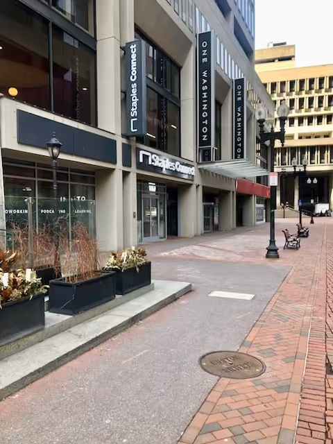 Urban outdoor sidewalk with storefronts, planters, and street lamps along a brick-paved pedestrian area.