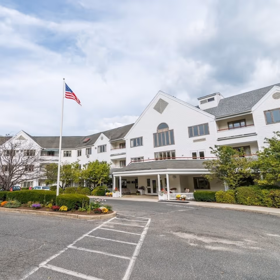 Front entrance of a white multi-story senior living building with an American flag, landscaped flower beds, and a parking area.