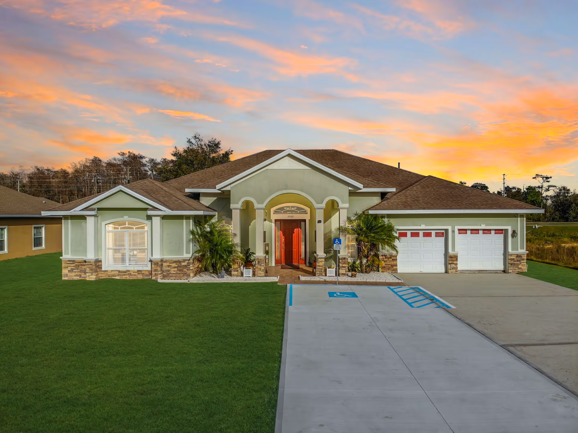 Single-story senior living building with a landscaped front entrance, garage doors, and a driveway with a marked wheelchair parking space under a colorful sunset.
