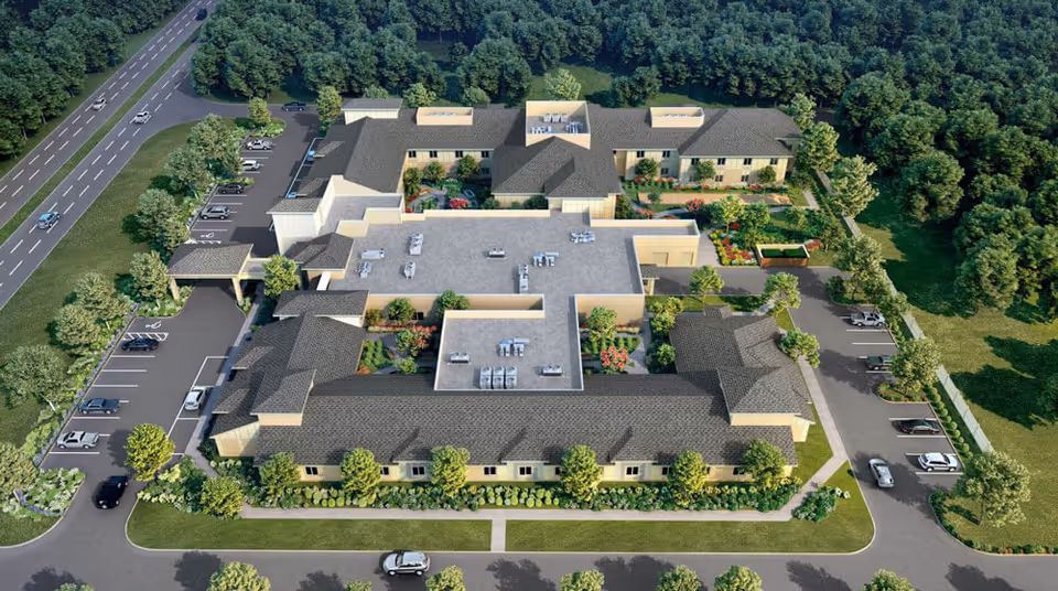 Aerial view of HearthStone at Leesburg senior living facility showing a large, single-story building with multiple wings, surrounded by landscaped gardens, parking lots with cars, and a road on one side. The building has a gray roof and beige walls, with green trees and bushes around the property.