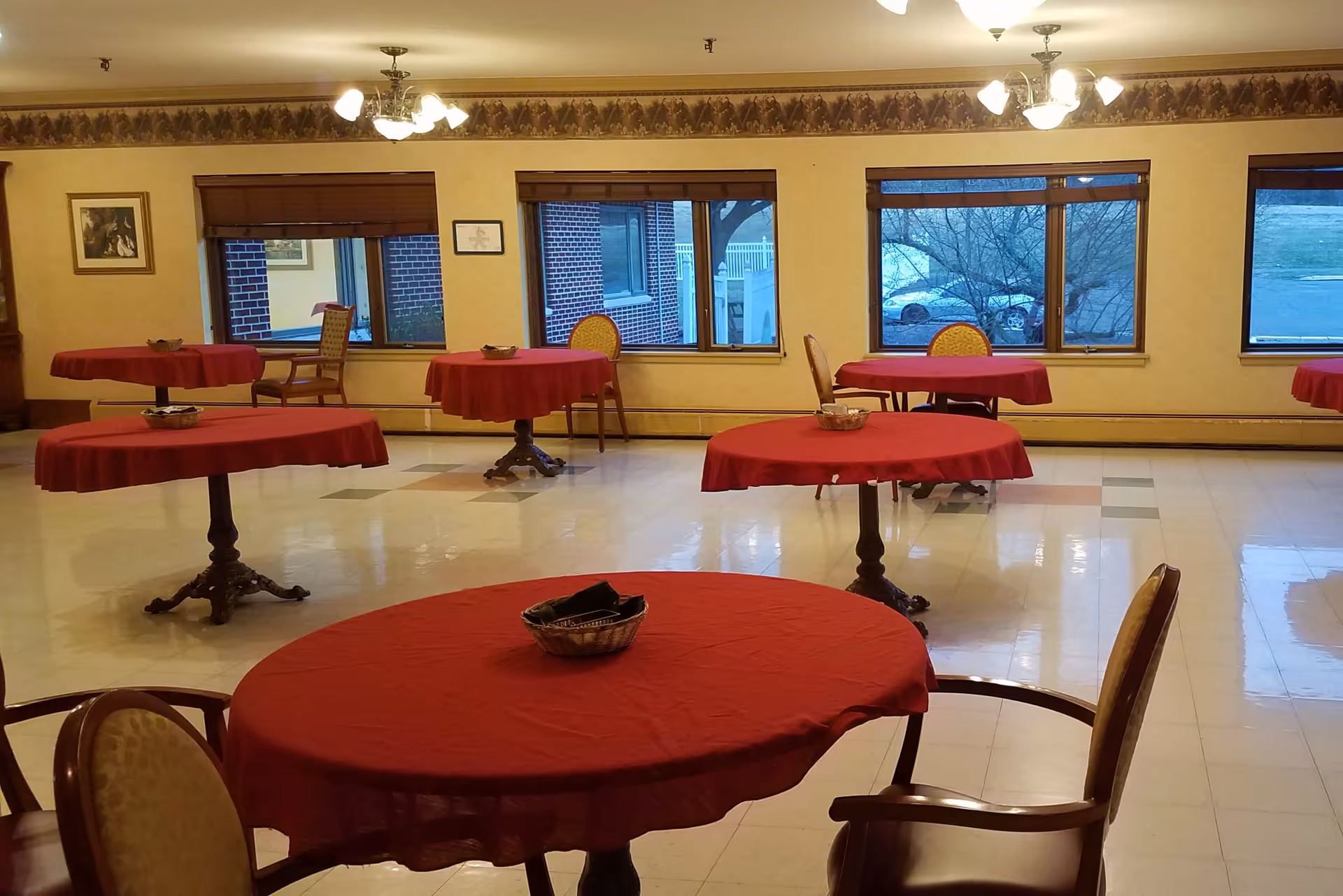 A dining room with several round tables covered with red tablecloths, each accompanied by chairs. The room has large windows showing an outdoor view with trees and a parked car. The walls are decorated with framed pictures and a floral border near the ceiling. Ceiling lights illuminate the space.