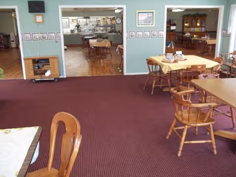 Empty dining room with wooden chairs, tables, and maroon carpeting in a senior living facility.