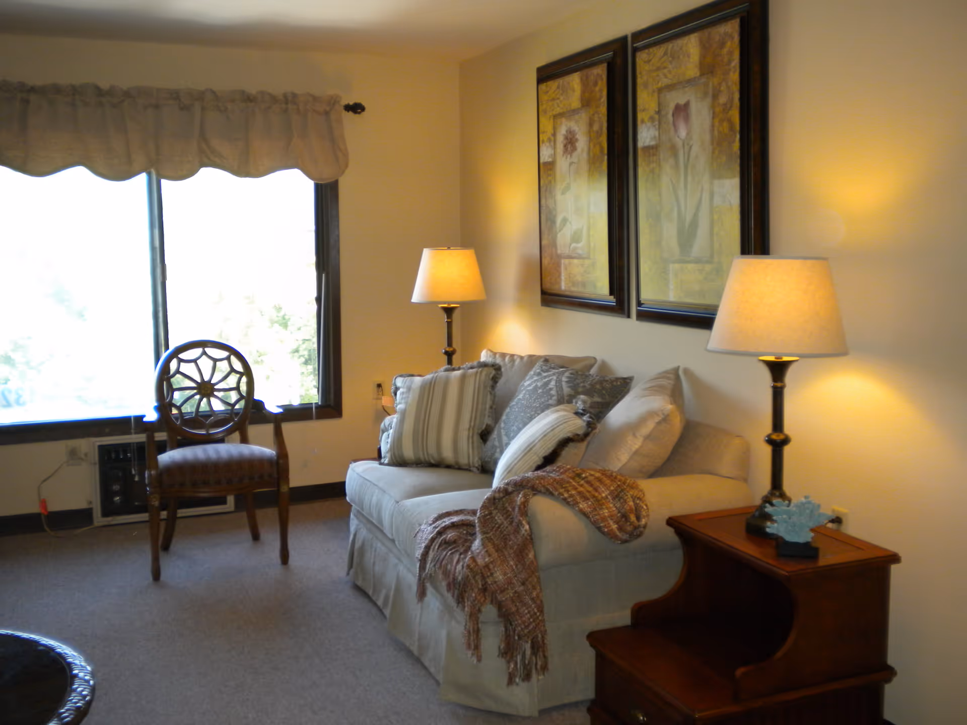 A cozy living room with a beige sofa adorned with multiple cushions and a throw blanket. Two framed floral paintings hang on the wall above the sofa. There are two table lamps with beige shades on either side of the sofa, one on a wooden side table. A wooden chair with a decorative backrest is positioned near a large window with a beige valance.