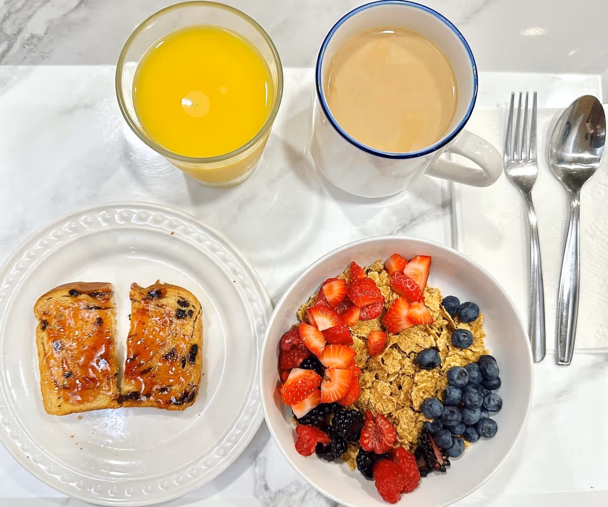 Top-down view of a meal: jam-topped toast, berry granola bowl, juice, and coffee on marble table.