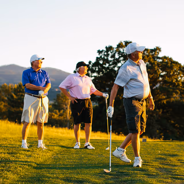 Three elderly men standing on a golf course holding golf clubs, with trees and hills in the background during sunset.