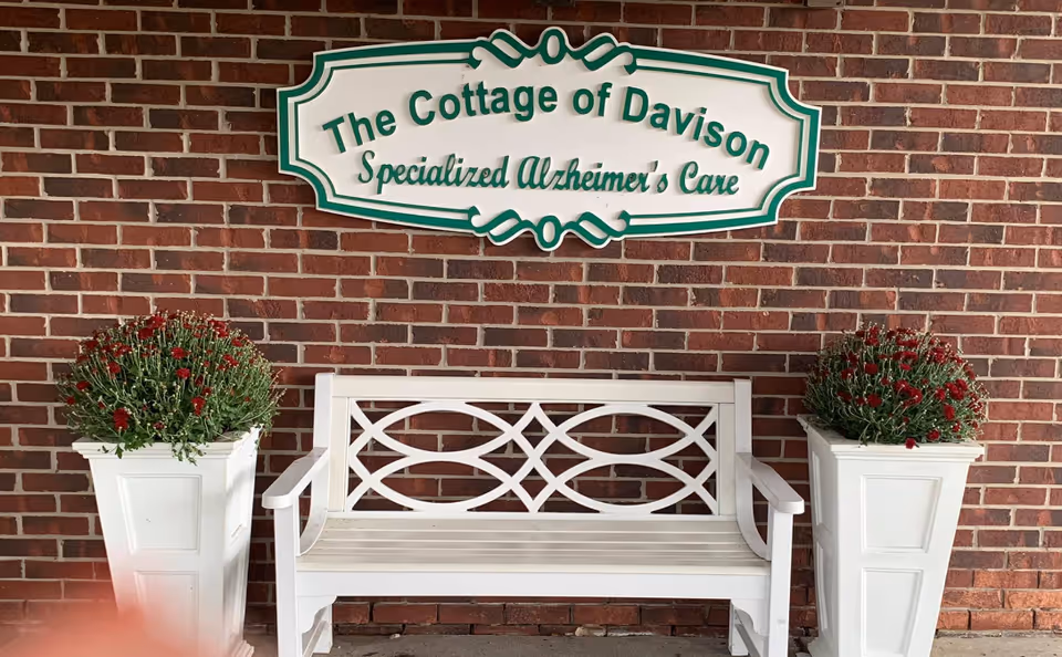 White bench between two planters with red flowers in front of The Cottage of Davison sign