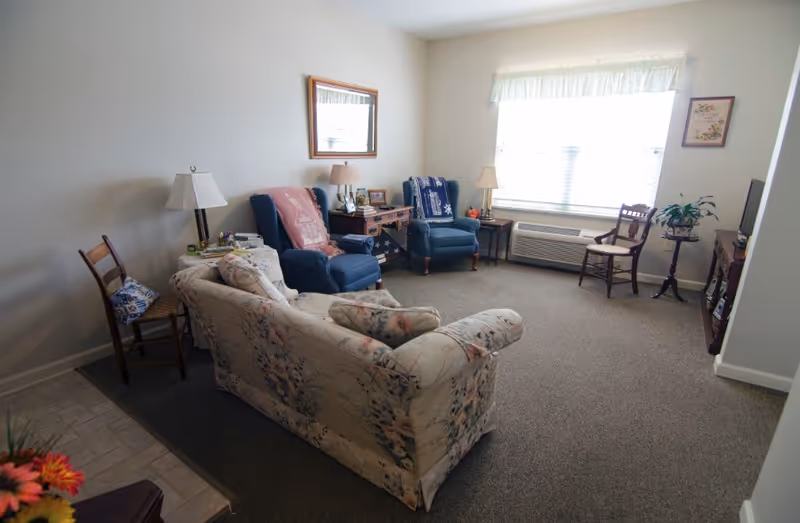 A cozy living room with a floral-patterned sofa, two blue armchairs with blankets draped over them, a wooden side table with a lamp and framed photos, a wooden chair, a small round table with a plant, a window with white blinds and a valance, and a television on a stand.