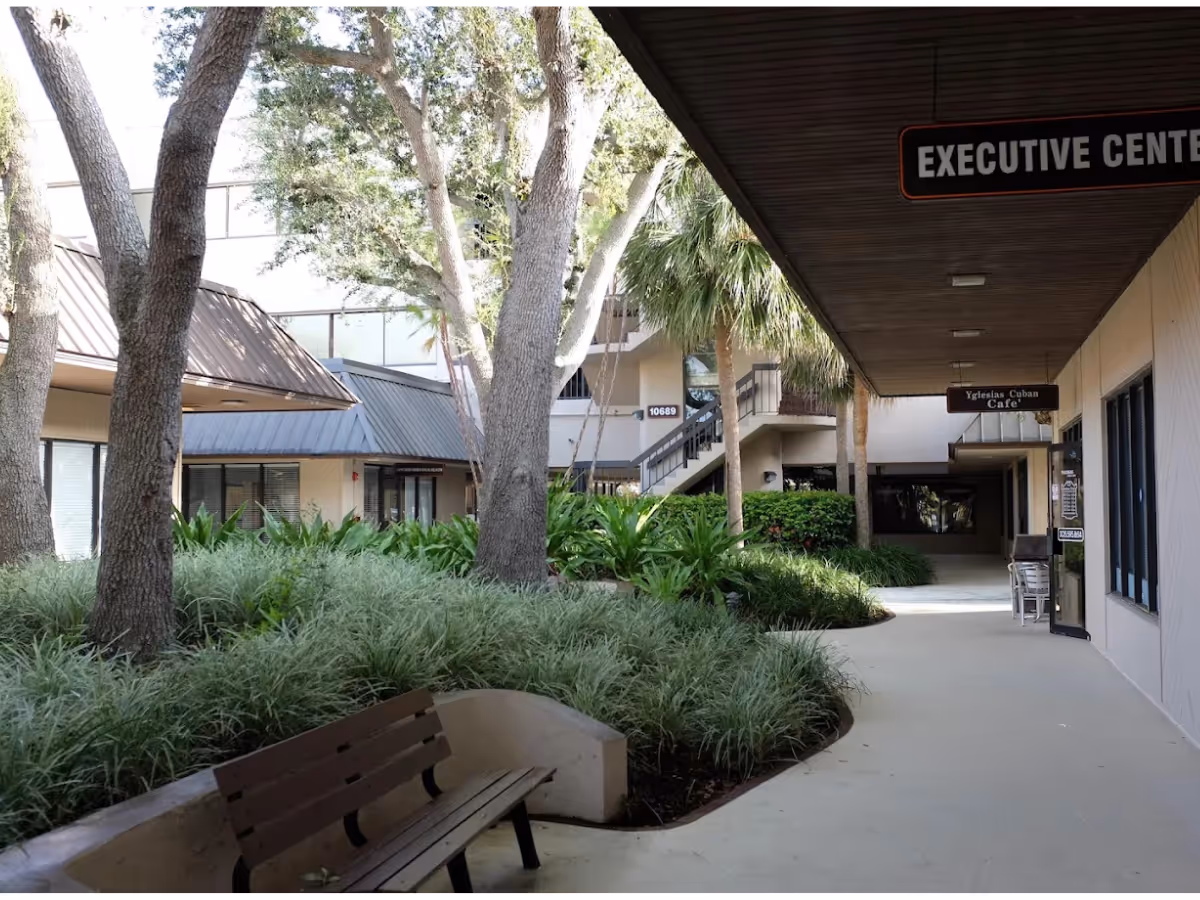 Outdoor walkway with trees, greenery, and benches outside a senior living facility.