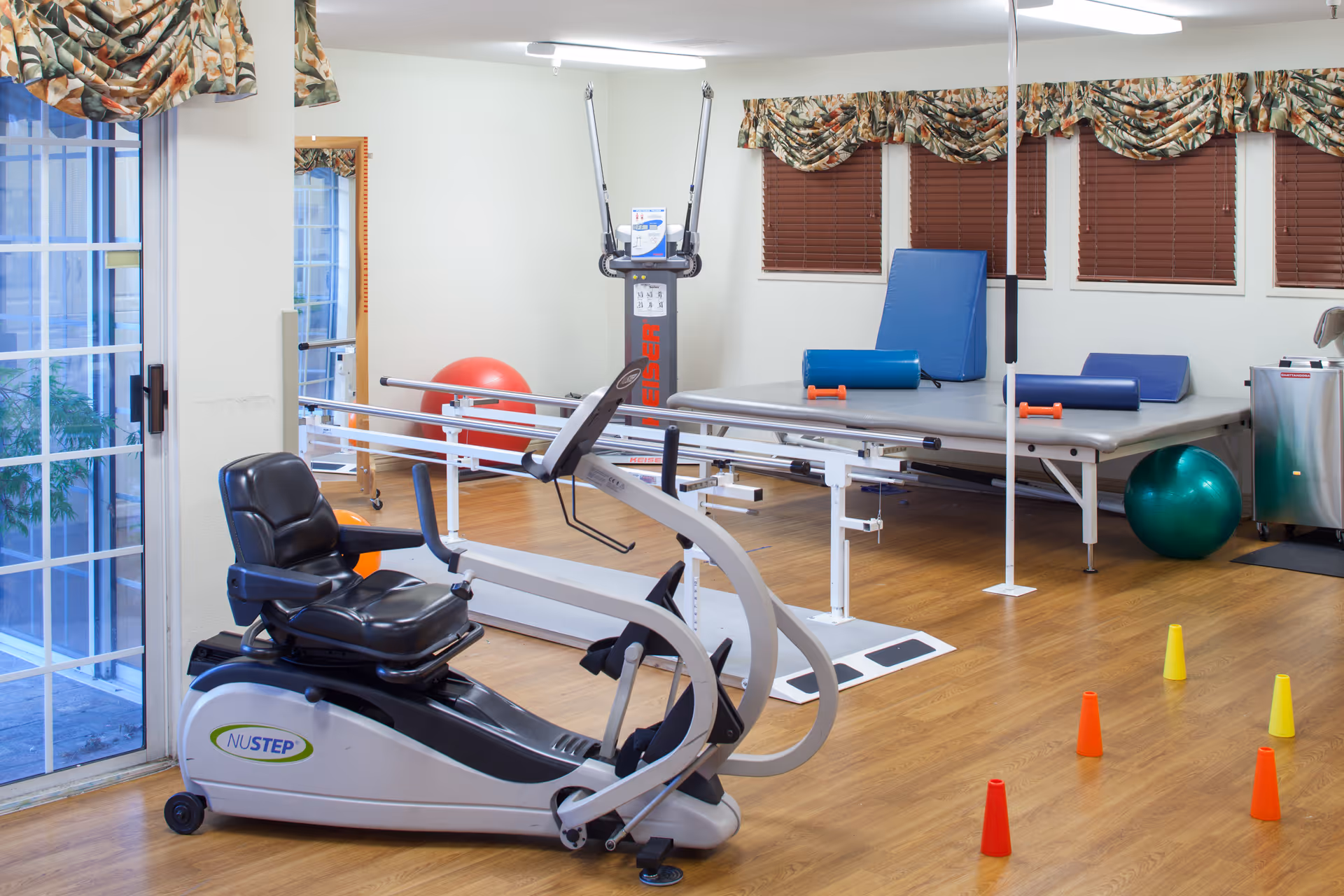 A physical therapy or rehabilitation room with exercise equipment including a NuStep recumbent cross trainer, parallel bars for walking support, therapy tables with foam rollers, exercise balls, and small colored cones on a wooden floor. The room has windows with floral valances and brown blinds, and a sliding glass door leading outside.