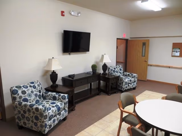 Senior living lounge with two patterned armchairs, lamps, a wall-mounted TV, and a round table.