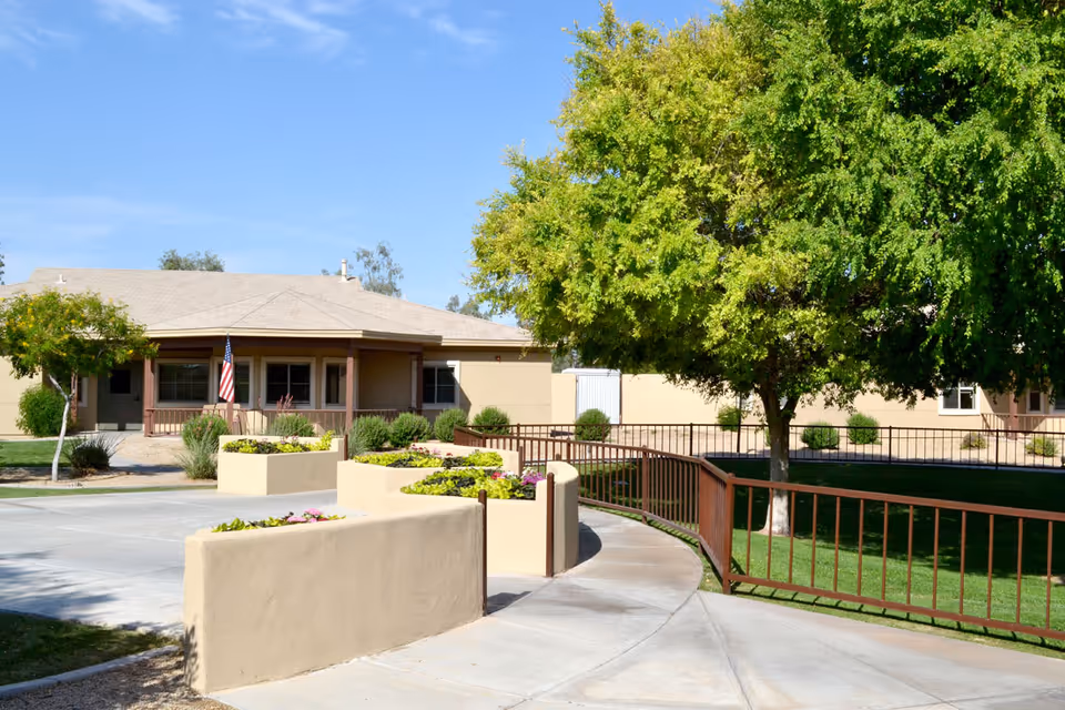 Front exterior of a single-story senior living building with a curved concrete walkway, raised planters, a large tree, and an American flag.