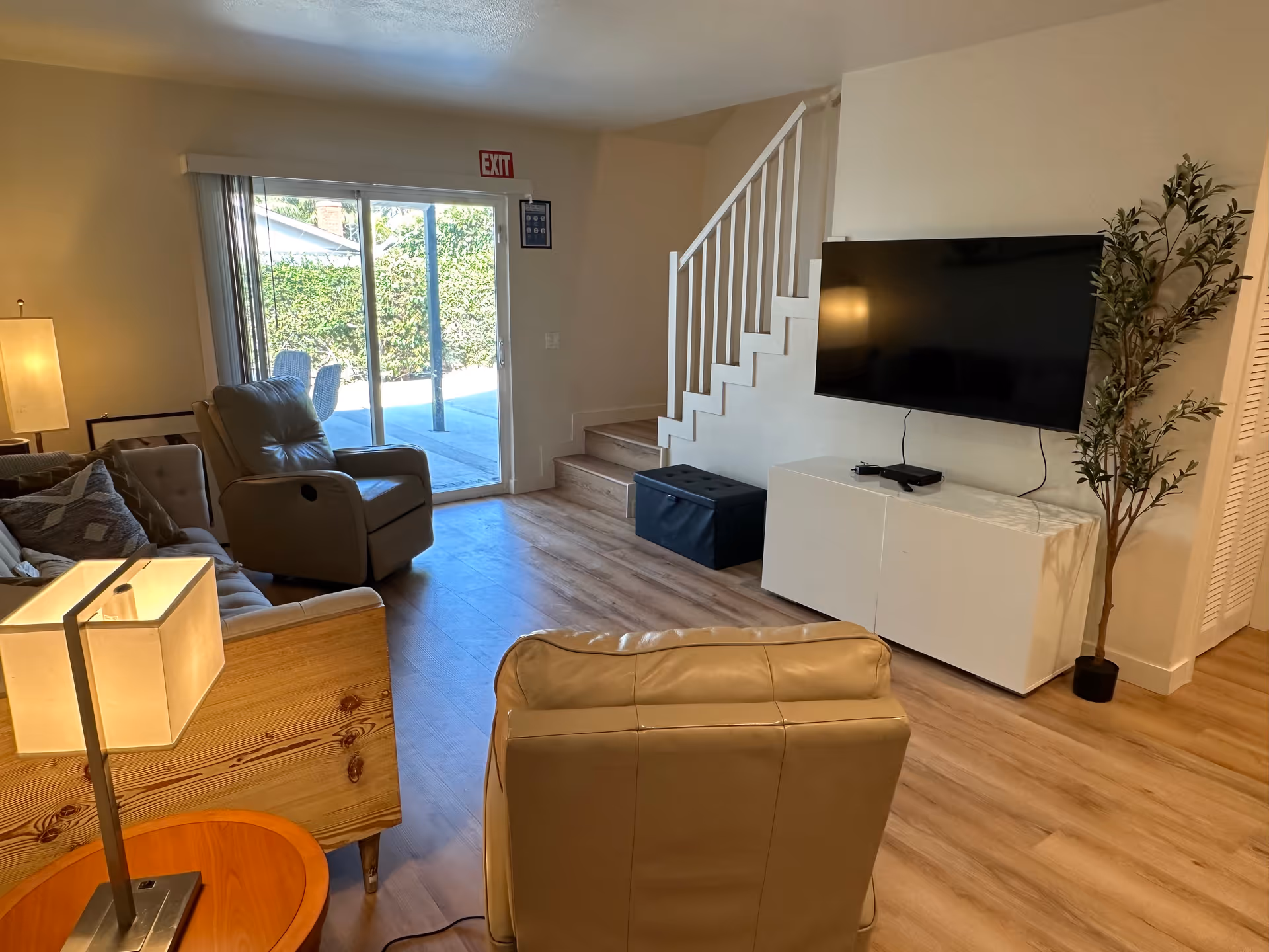 A cozy living room with light wood flooring, a beige leather recliner chair, a sofa with cushions, a wooden side table with a lamp, a flat-screen TV mounted on the wall above a white cabinet, a potted plant in the corner, and sliding glass doors leading to an outdoor patio area.