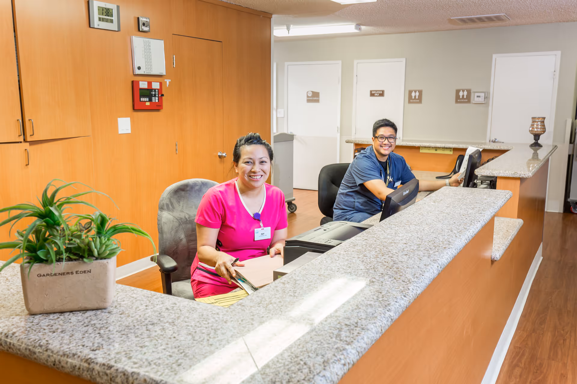 Two smiling healthcare workers sitting behind a reception desk in a medical facility. One is wearing a pink uniform and the other is in a blue uniform. The desk has a granite countertop and there is a potted plant on it. Behind them are wooden cabinets and doors with signs indicating staff-only areas and restrooms.