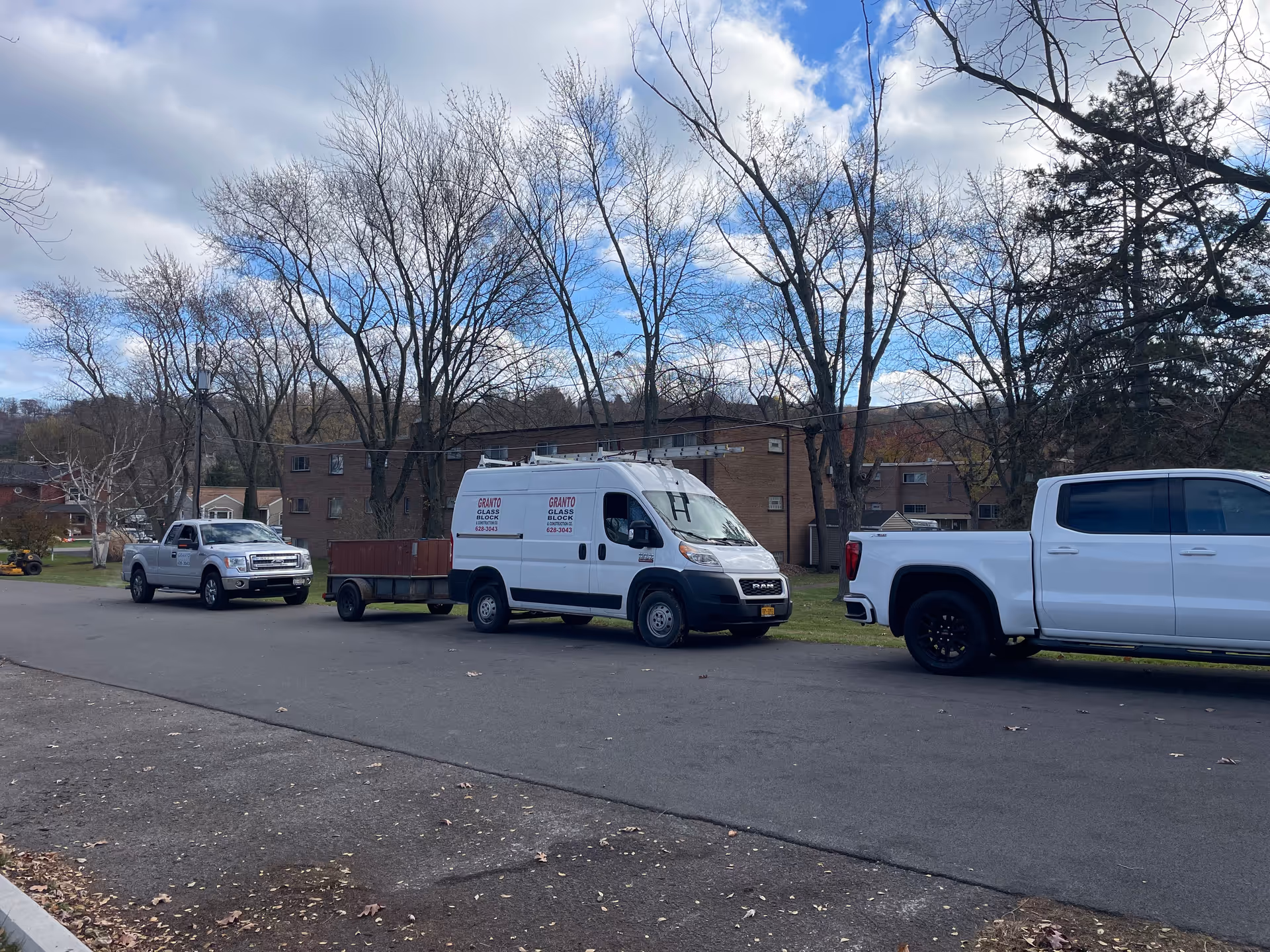 Exterior street view with trees, a brick building, and parked white service vehicles.