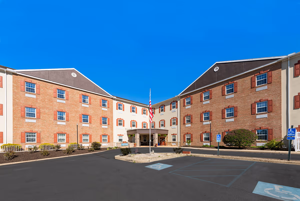 Brick senior living facility with central entrance, flagpole, and parking lot under a clear blue sky