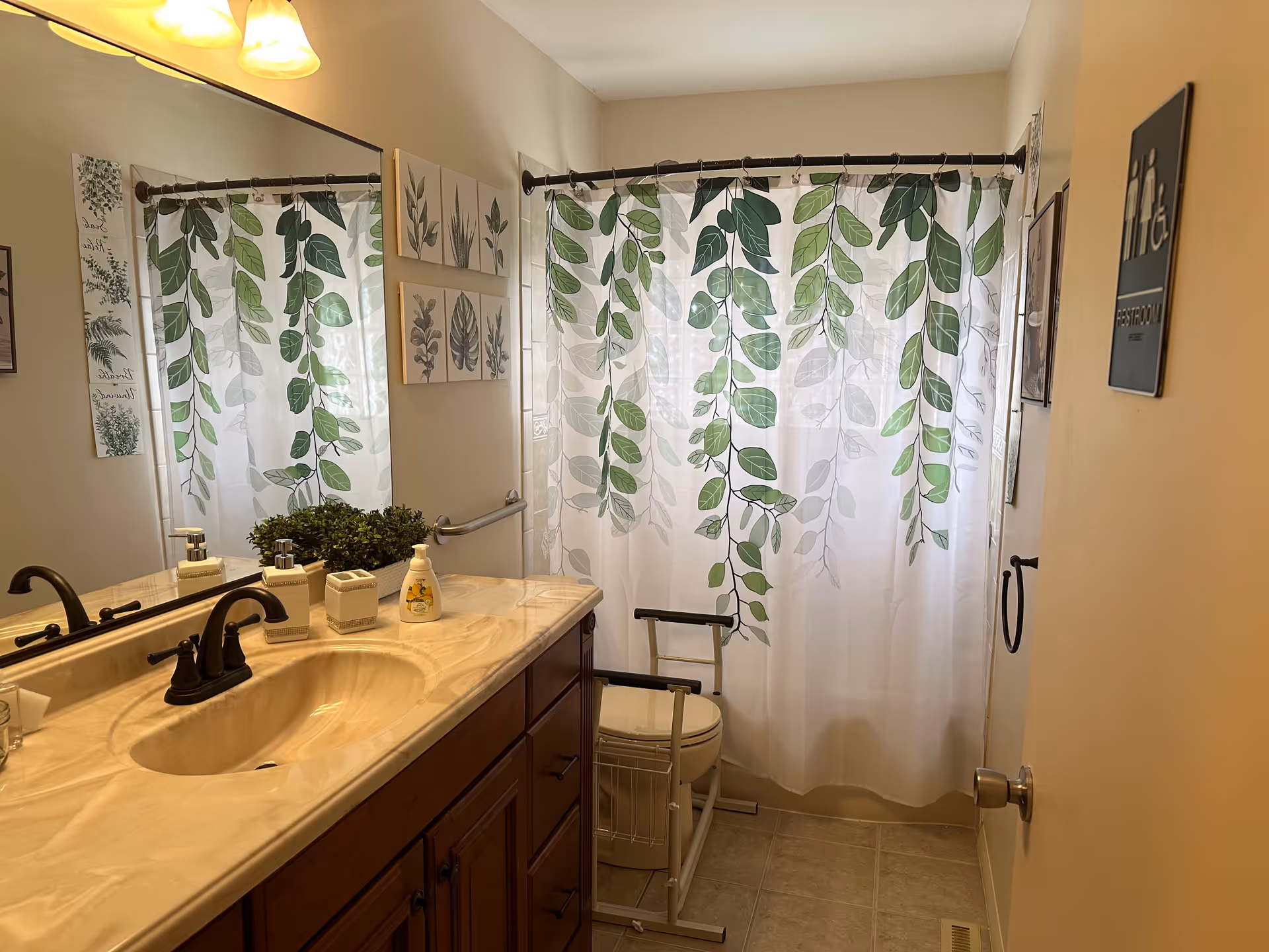 Well-lit bathroom with a sink and vanity, a leafy-pattern shower curtain, mirror, and a raised toilet frame.