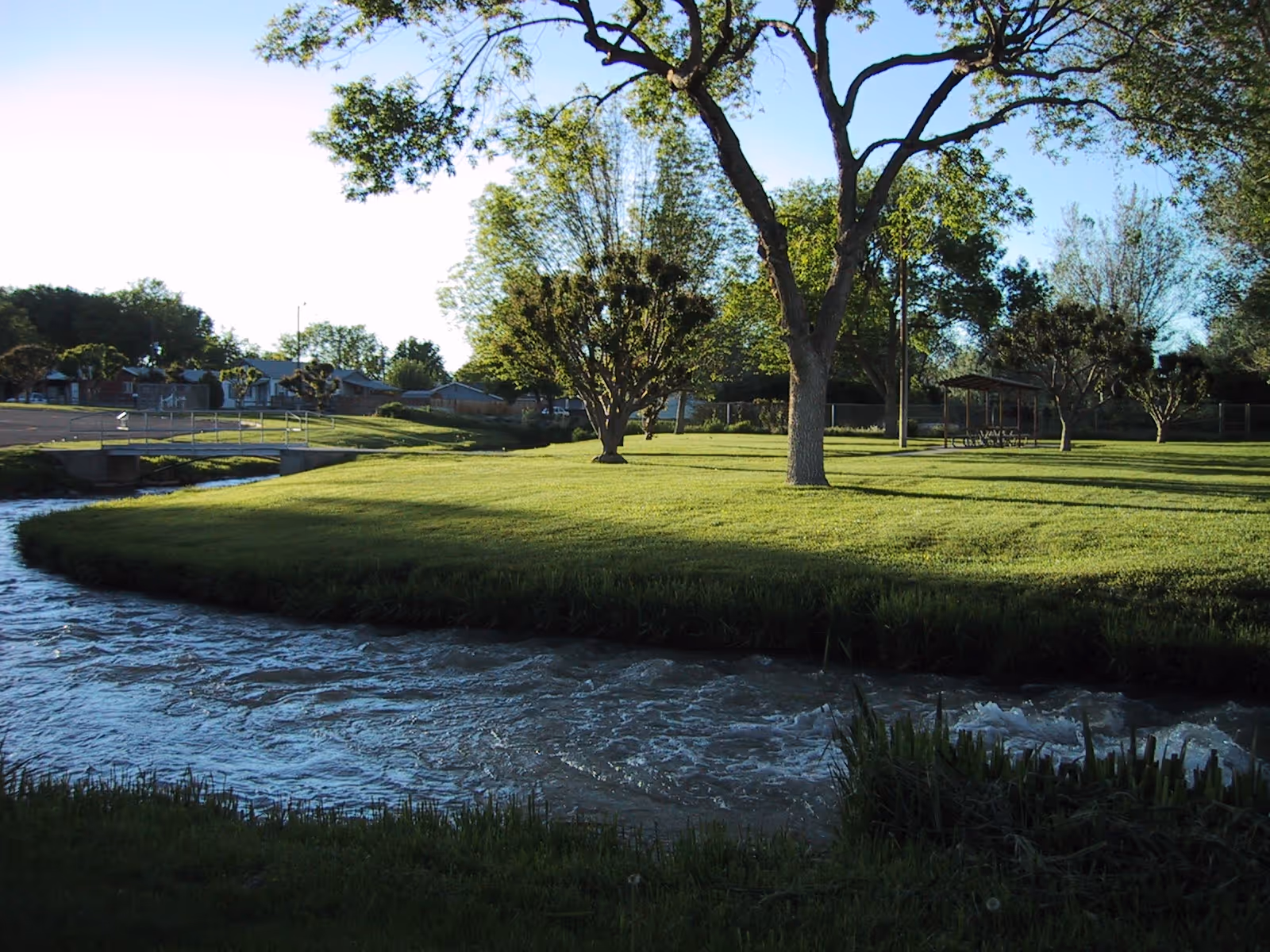 Sunlit park-like outdoor area with grassy lawn, trees, a river, and a shaded pavilion.