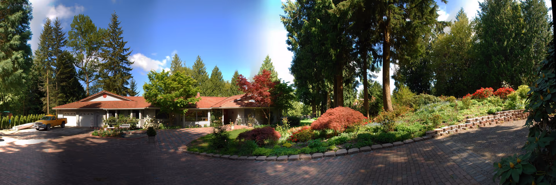 Panoramic view of a single-story residential building with a circular brick driveway and landscaped gardens surrounded by tall trees.