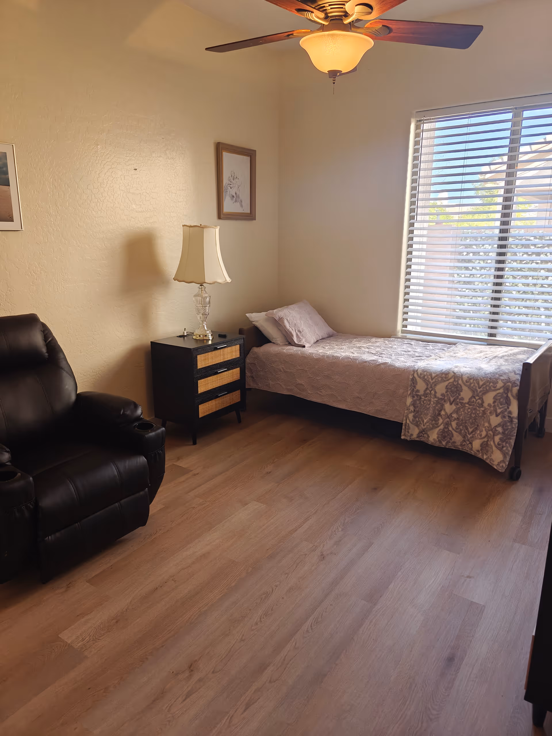 A furnished bedroom with a bed, nightstand, lamp, recliner, and large window blinds.