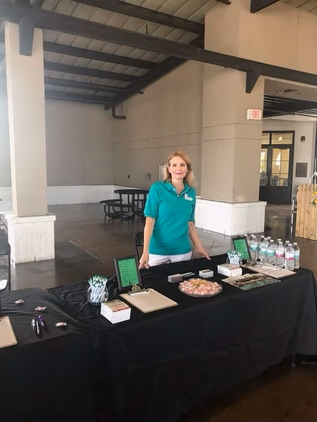 Staff member in teal polo stands behind a promotional table in a large indoor hall.