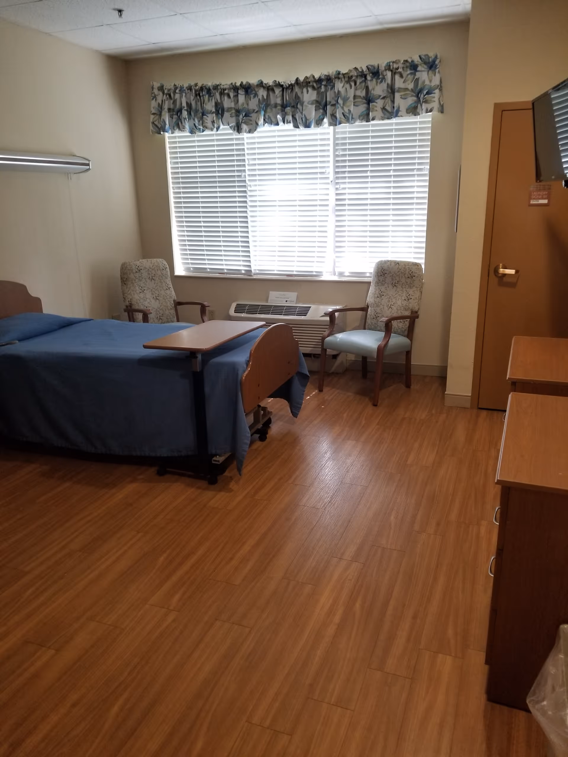 A patient room in Palm Garden of Largo with a bed, two chairs, and window blinds.