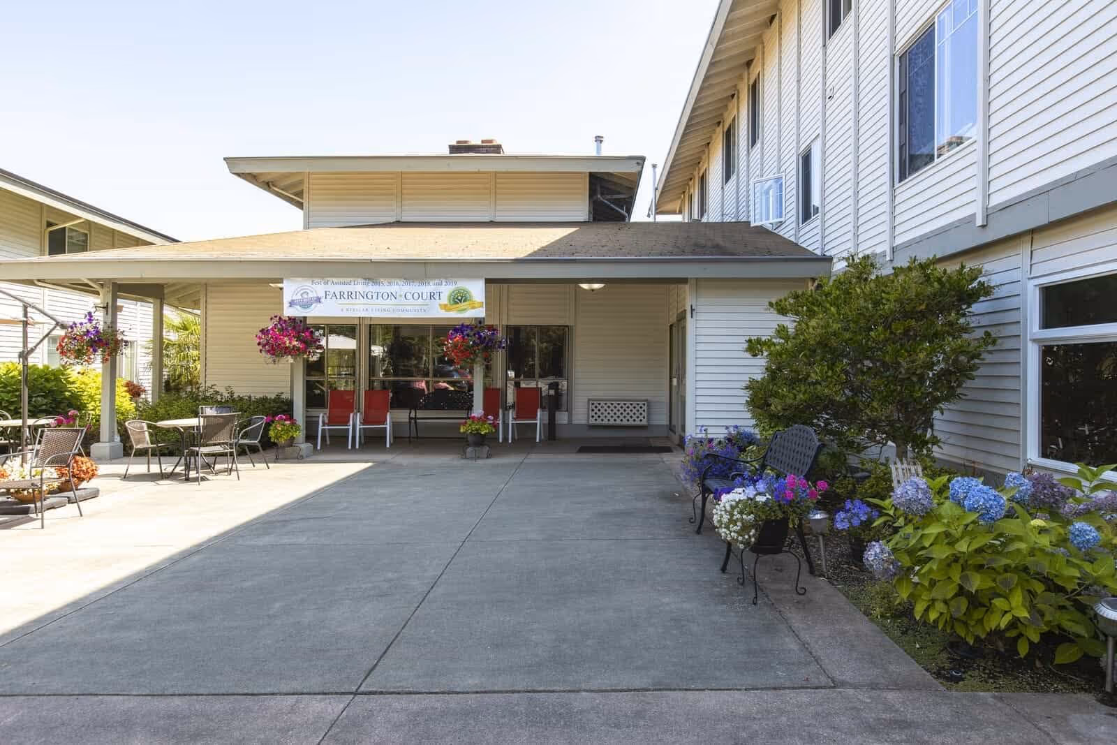 Front entrance of a retirement home with a covered porch, outdoor seating, hanging flower baskets, and potted flowers along the walkway.