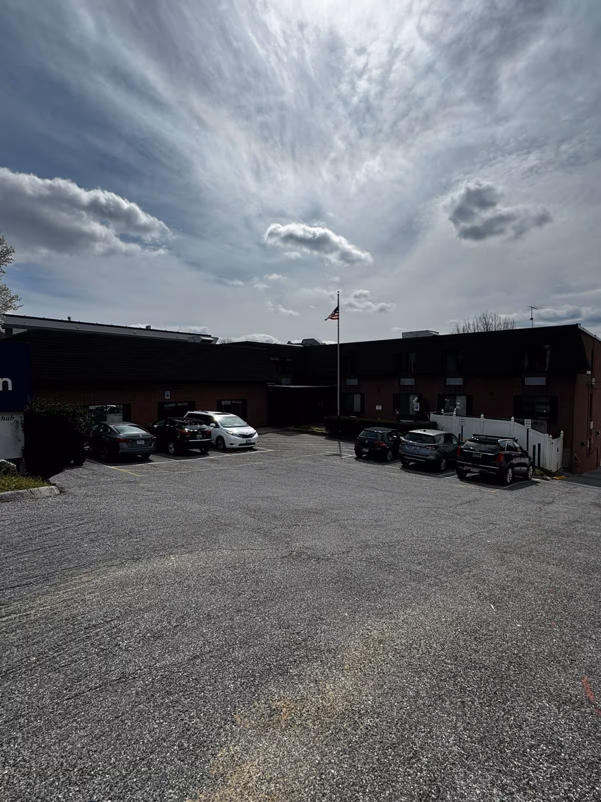 Parking lot in front of a two-story brick building with several parked cars and an American flag on a flagpole under a partly cloudy sky.