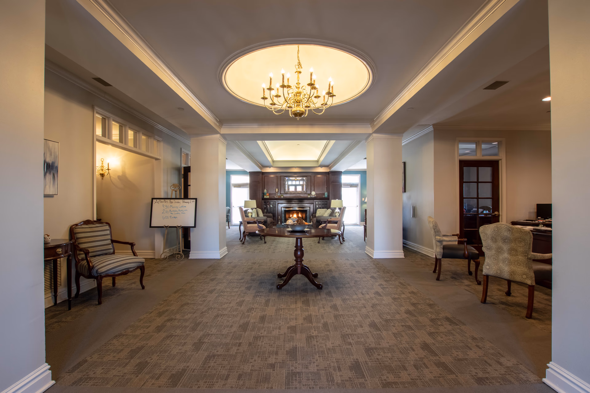 A spacious and elegant common area in a senior living facility with a central round wooden table, a chandelier hanging from a recessed ceiling, and a cozy fireplace surrounded by armchairs and sofas in the background. The room has carpeted floors, neutral-colored walls, and additional seating areas on both sides.