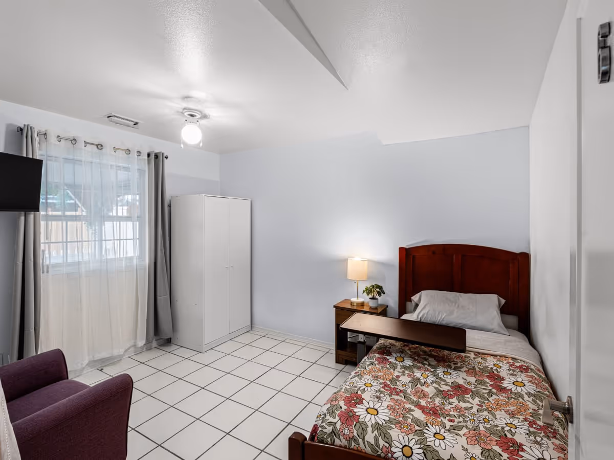 A single bedroom with bed, wardrobe, nightstand, lamp, chair, and a window.