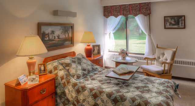 A cozy bedroom in a senior living facility with a single bed covered by a patterned quilt. There are two bedside tables with lamps and framed photos. A window with sheer white curtains and a valance lets in natural light, and a cushioned wooden rocking chair sits near a small round table with a flower arrangement and an open book on a tray.