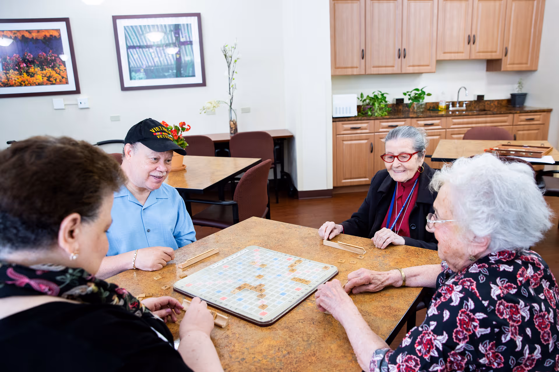 Four elderly individuals sitting around a table playing a word tile board game in a room with wooden cabinets and framed pictures on the wall.