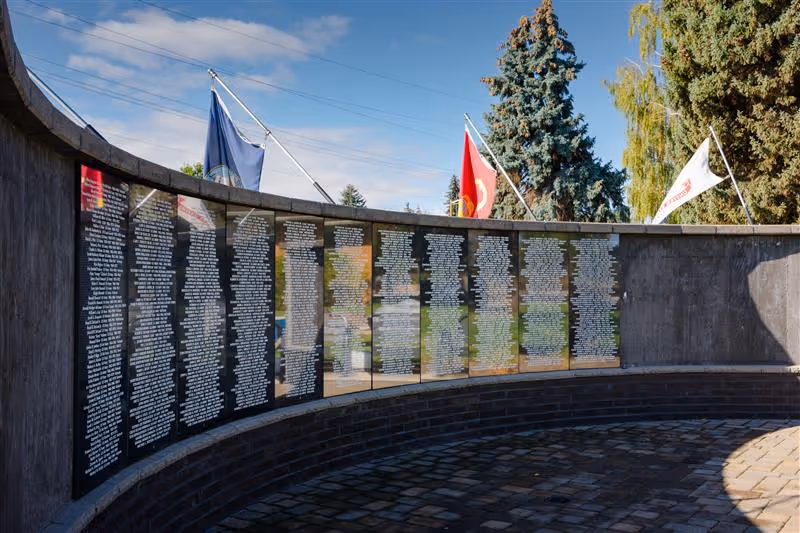 Curved outdoor memorial wall with vertical plaques, flags, and surrounding trees.