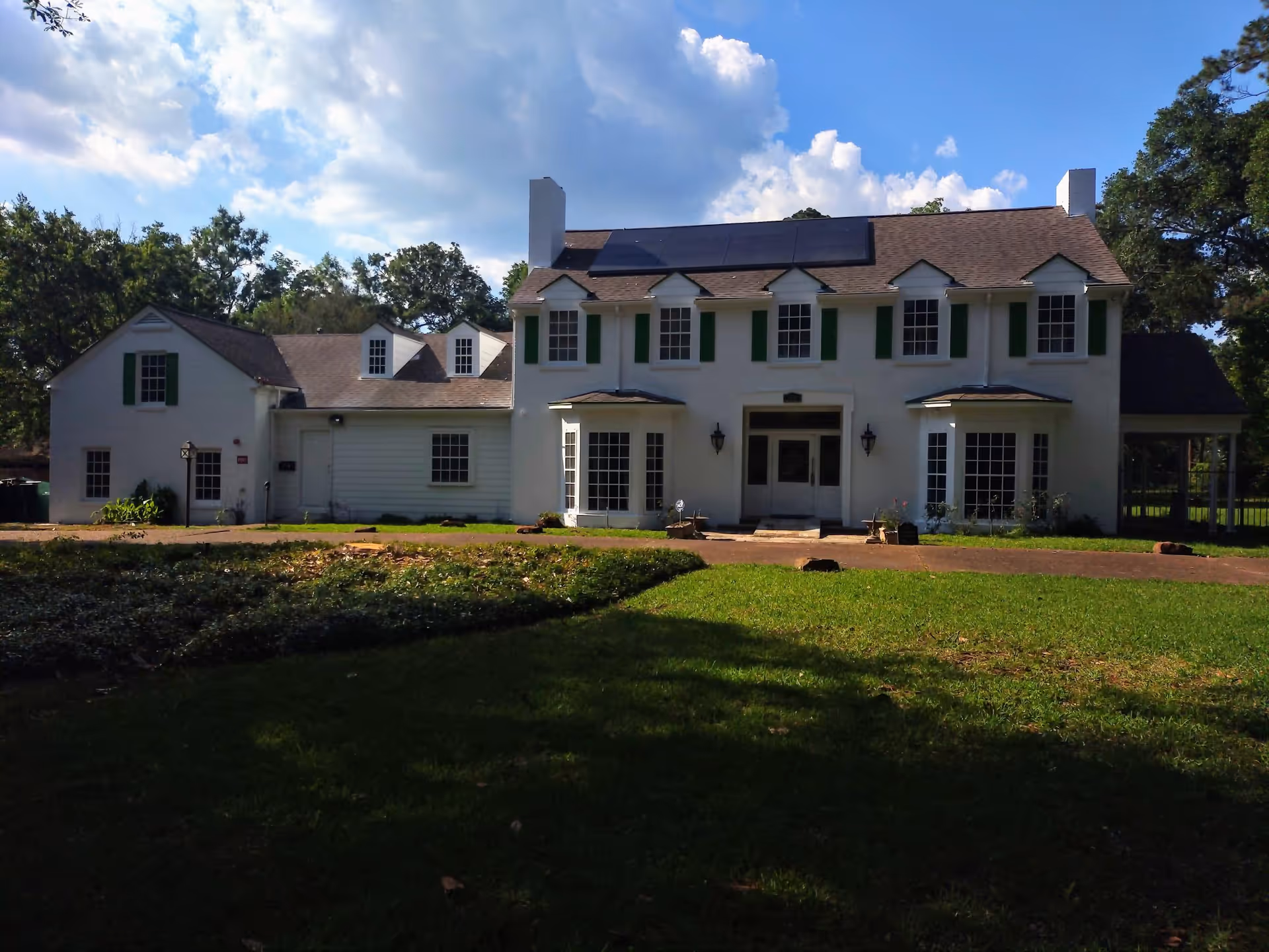 Two-story white assisted living building with green shutters, multiple windows and a driveway in front under a partly cloudy sky.