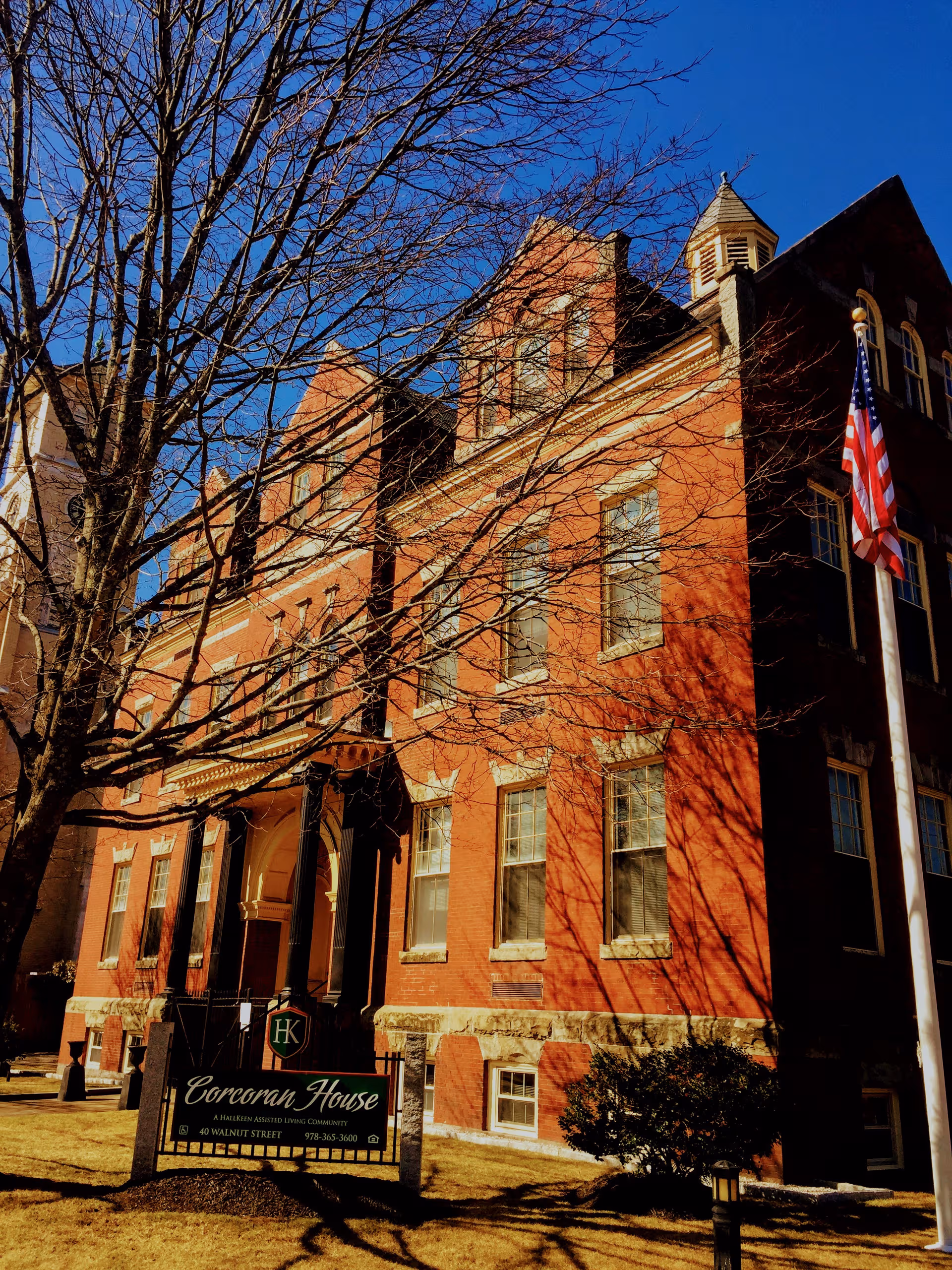 Front exterior of Corcoran House assisted living, red brick building with flag under blue sky.