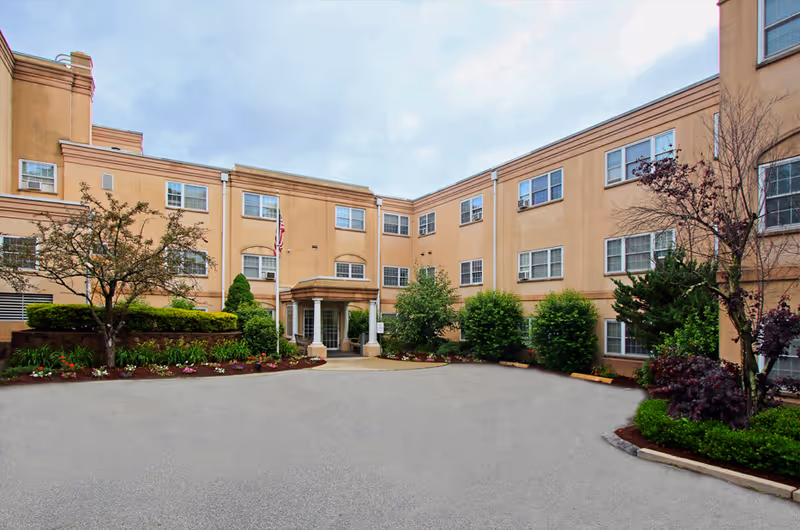 Front entrance and driveway of a three-story beige senior living facility with landscaping and a flagpole.