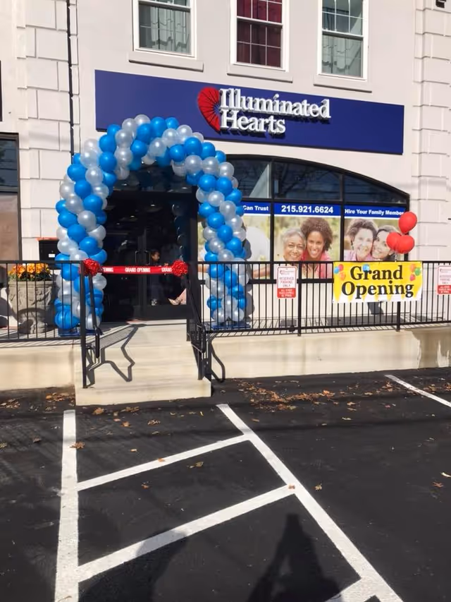 Front entrance of Illuminated Hearts senior living facility with blue balloon arch and Grand Opening banner.