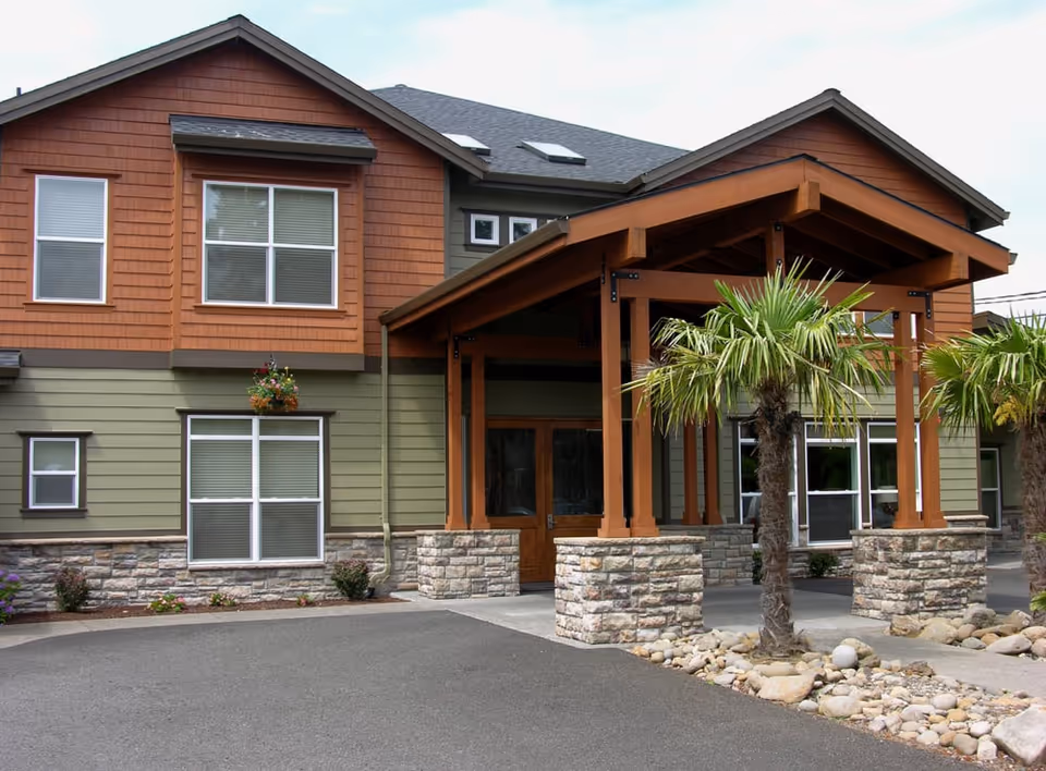 Entrance of a two-story building with a covered wooden porte-cochere, stone pillars, palm trees, and multiple windows.